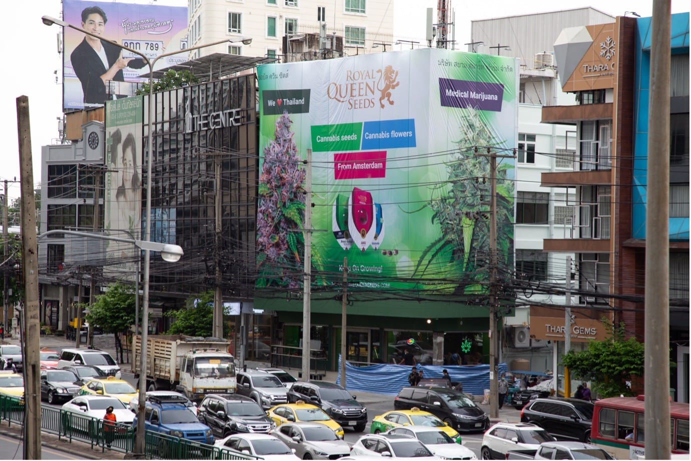 Busy Bangkok street with heavy traffic passing a cannabis dispensary beneath a large Royal Queen Seeds billboard.
